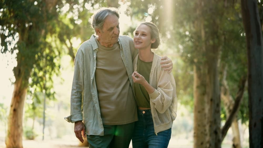 An elderly father and his daughter walking arm in arm through sunlit woodland